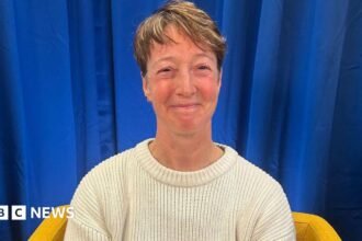 Nicky Perfect looks at the camera and smiles. She is wearing a cream wool jumper and sits on a yellow armchair. The backdrop is a blue curtain