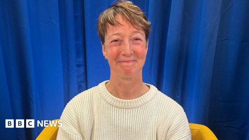 Nicky Perfect looks at the camera and smiles. She is wearing a cream wool jumper and sits on a yellow armchair. The backdrop is a blue curtain