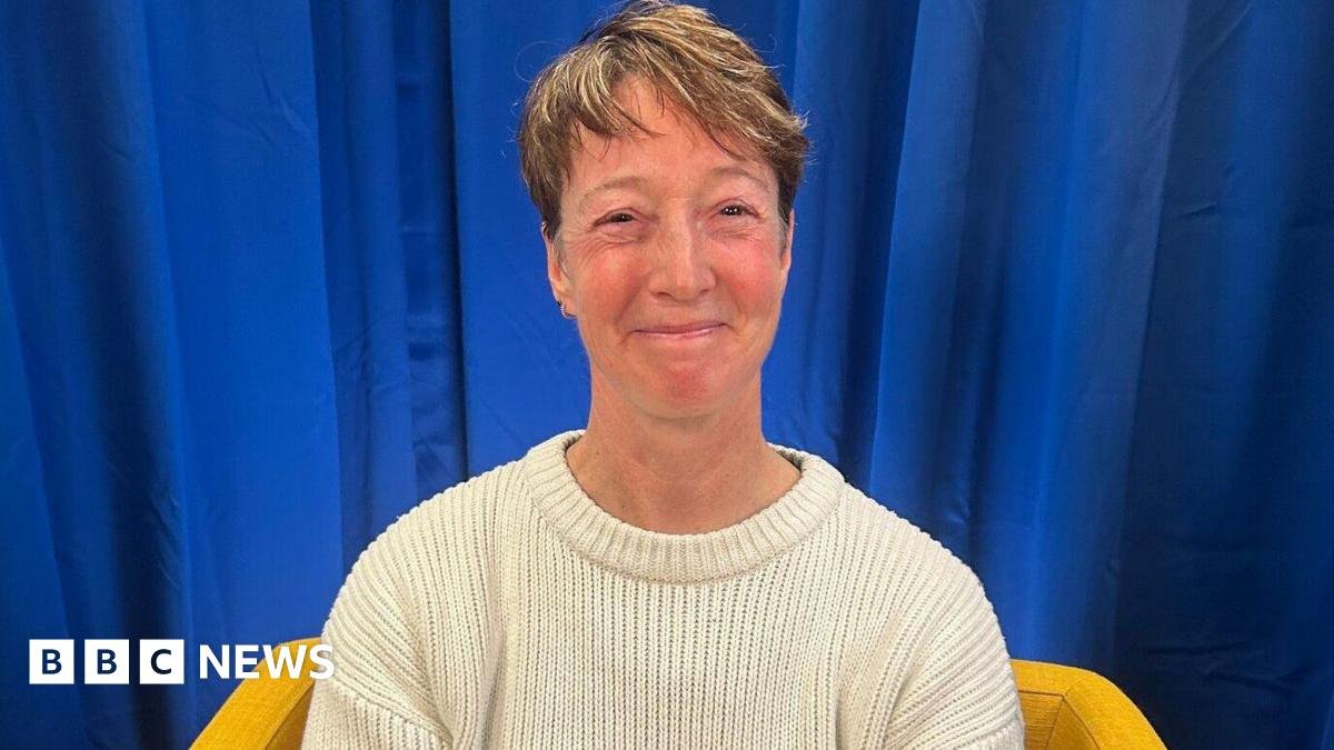 Nicky Perfect looks at the camera and smiles. She is wearing a cream wool jumper and sits on a yellow armchair. The backdrop is a blue curtain