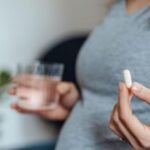 A pregnant women wearing a grey top is visible from the chest down, and holds a white pill in her left hand and a glass of water in her right (slightly blurred)