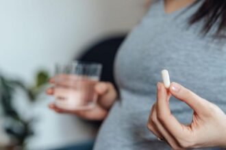 A pregnant women wearing a grey top is visible from the chest down, and holds a white pill in her left hand and a glass of water in her right (slightly blurred)