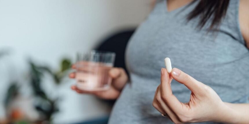 A pregnant women wearing a grey top is visible from the chest down, and holds a white pill in her left hand and a glass of water in her right (slightly blurred)