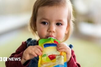 A toddler with blondey-brown hair sucking a pouch of baby food, which she is holding with both hands. She is wearing a burgundy top and dungarees.