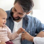A baby smiling and sitting with her parents, alongside their pediatrician who is touching the baby