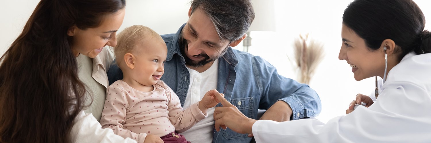 A baby smiling and sitting with her parents, alongside their pediatrician who is touching the baby